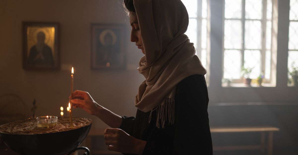 A woman in a headscarf lights a candle in a sunlit church, surrounded by religious icons.